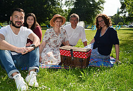 Menschen haben sich mit Picknick-Korb und Decke auf dem Rasen im Park niedergelassen und lächeln in die Kamera