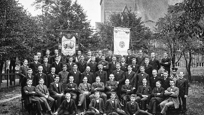 Schwarzesweißes Gruppenfoto mit ca. 30 Männern, im Hintergrund die beiden Fahnen und die Kirche in Neumünster