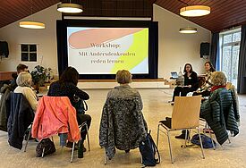Menschen sitzen bei einem Workshop im Stuhlkreis und schauen auf einen Monitor