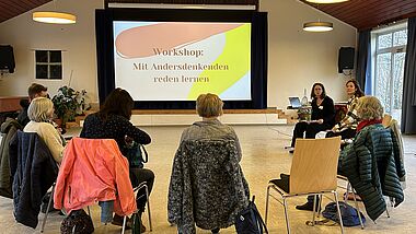 Menschen sitzen bei einem Workshop im Stuhlkreis und schauen auf einen Monitor