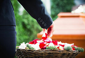 Eine Hand, die in einen Korb mit Blütenblättern greift, im Hintergrund ist ein Sarg zu sehen