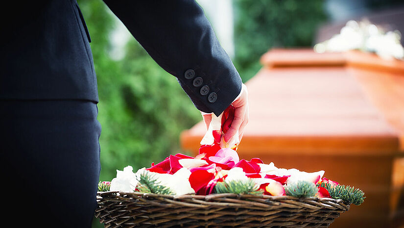Eine Hand, die in einen Korb mit Blütenblättern greift, im Hintergrund ist ein Sarg zu sehen