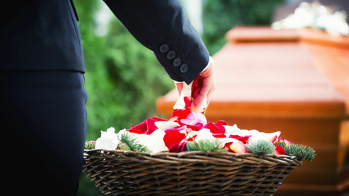 Eine Hand, die in einen Korb mit Blütenblättern greift, im Hintergrund ist ein Sarg zu sehen