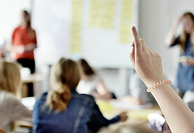 Im Vordergrund: Eine Hand, die sich meldet, im Hintergrund unscharf: Schüler, die an Tischen sitzen, eine Tafel, davor zwei Lehrpersonen