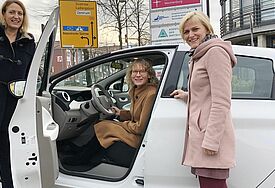 Weißes E-Auto, Tür geöffnet, darin sitzt eine Frau, daneben stehen zwei weitere und lächeln.