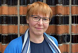 Eine Frau mir kurzen rotblonden Haaren und Brille blickt freundlich lächelnd in die Kamera. Sie trägt eine hellblaue Strickjacke, um ihren Hals hängt ein blau-Weiß-gemustertes Tuch..