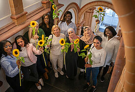 Frauen multiethnischer Herkunft stehen in einem Treppenhaus und halten strahlend Sonnenblumen in die Höhe.