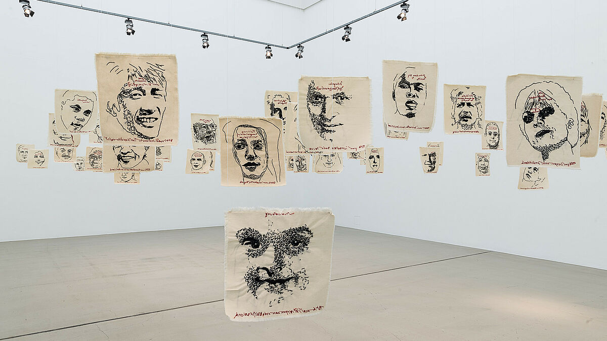 Das Kunstwerk Einwanderung von Künstlerin Sybille Loew:56 auf Leinwand gestickte Portraits von Menschen mit Migrationshintergrund hängen von der Decke der Galerie. Auf Augenhöhe montiert, man sieht sie von Angesicht zu Angesicht.