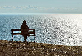 Eine Frau sitzt alleine auf einer Bank an einer Steilküste. Von ihr ist nur eine dunkle Silhouette zu sehen, das Meer glitzert in der Sonne.