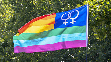 Bunte Regenbogenfahne mit doppeltem Frauensymbol vor grüner Baumkrone
