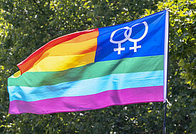 Bunte Regenbogenfahne mit doppeltem Frauensymbol vor grüner Baumkrone