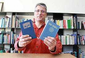 Mann in rotem Pullover hält eine Bibel und einen Koran in der Hand, im Hintergrund ein Regal mit vielen Büchern.