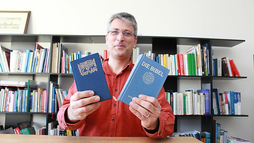 Er ist der Islambeauftragte der Nordkirche: Sönke Lorberg-Fehring. Mann in rotem Pullover hält eine Bibel und einen Koran in der Hand, im Hintergrund ein Regal mit vielen Büchern.