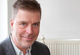 Porträt von Henning Kiene, er trägt Hemd und Sakko, lächelt in die Kamera