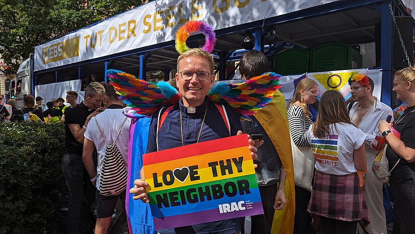 Pastor mit Schild in Regenbogenfarben und Schriftzug: "Love Thy Neighbor"