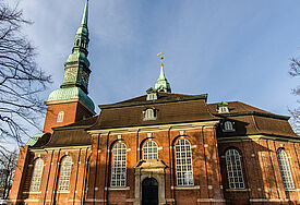 Kirche im barocken Stil mit grün bedachtem Kirchturm, Himmel ist blau, Rasen davor grün, umgeben von laublosen Bäumen