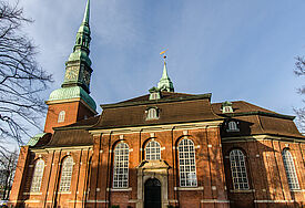 Kirche im barocken Stil mit grün bedachtem Kirchturm, Himmel ist blau, Rasen davor grün, umgeben von laublosen Bäumen