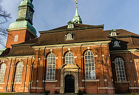 Kirche im barocken Stil mit grün bedachtem Kirchturm, Himmel ist blau, Rasen davor grün, umgeben von laublosen Bäumen
