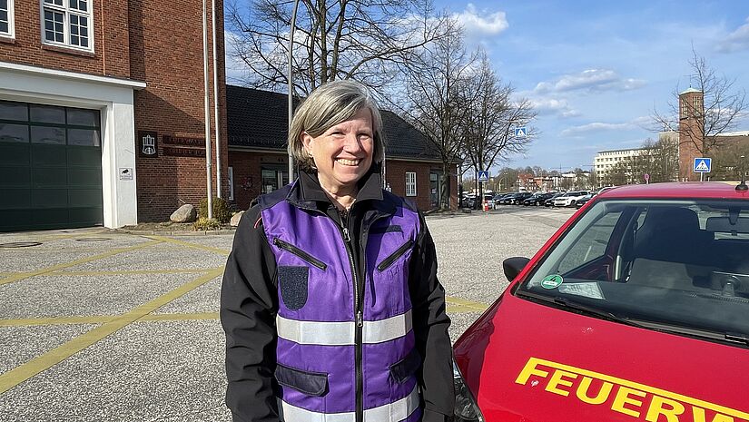 Eine Frau mit einer violetten Weste steht neben einem kleinen roten Auto mit der Aufschrift Feuerwehr