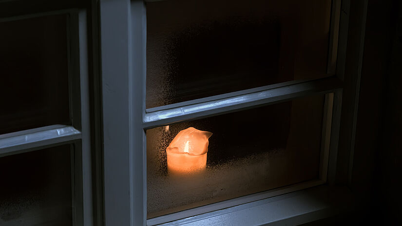Eine Kerze brennt hinter einer beschlagenen Glasscheibe.