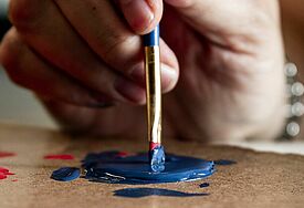 Eine Hand tunkt einen Pinsel in blaue Farbe.