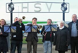 Eine Gruppe an Menschen steht mit Instrumenten und Notenständern vor dem Stadion, über dem groß "HSV" prangt