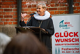 Bischöfin Kirsten Fehrs spricht vor der Mauer des Dom zu Lübeck am Stehpult ihren Abendsegen anlässlich des 850. Geburtstags des Dom, hinter ihr ein Plakat mit dem Aufdruck: Glückwunsch Geburtstagskind 