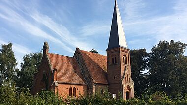 Die Dorfkirche aus rotem Backstein in Lüssow steht umgeben von grünen Bäumen unter blauem Himmel.