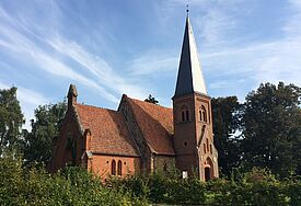 Die Dorfkirche aus rotem Backstein in Lüssow steht umgeben von grünen Bäumen unter blauem Himmel.