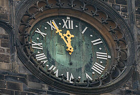 Die goldenen Zeiger einer Turmuhr stehen auf fünf vor 12, der Hintergrund der Uhr schimmert grünlich, die Steine des Turm sind graubraun..