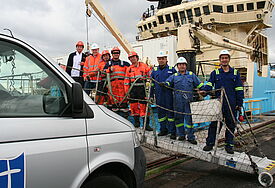 Im Vordergrund ein Fahrzeug der Seemannsmission, schräg daneben Seefahrer, dahinter ein Frachter