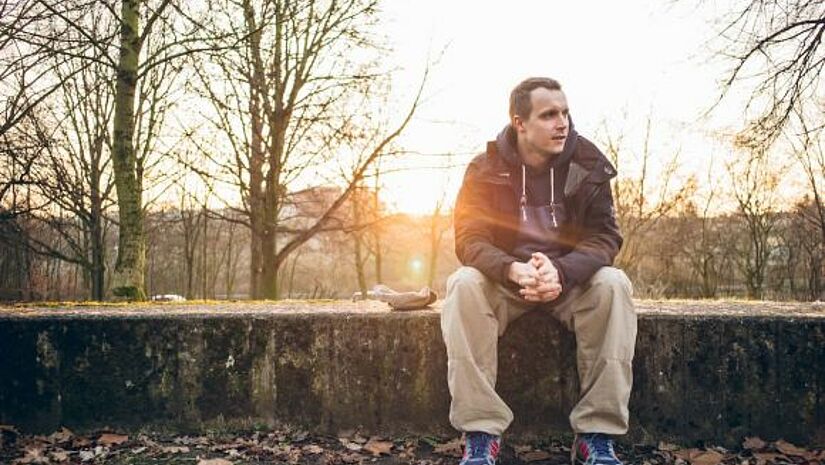 Martin Fehrensen sitzt auf einer niedrigen Steinwand und schaut nach rechts, im Hintergrund leuchtet zwischen drei Bäumen die Sonne