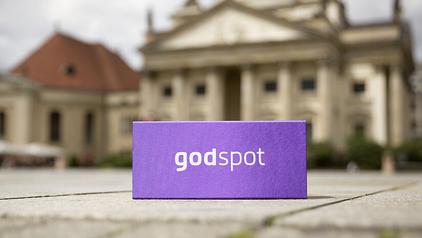 Lila Godspot-Karton draußen auf dem Weg - dahinter ein kirchliches Gebäude