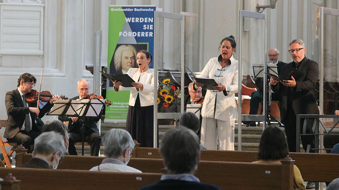 Sängerinnen, Sänger und Musiker stehen im Dom zu Greifswald und musizieren.  Aufgrund der Corona-Pandemie stehen Scheiben aus Plexiglas zwischen den Singenden.