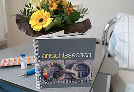 Das Buch im quadratischen Format steht auf einem kleinen Beitisch neben einem Krankenhausbett, im Hintergrund ein Strauß Blumen, ein Kugelschreiber und eine Blisterpackung mit Medikamenten