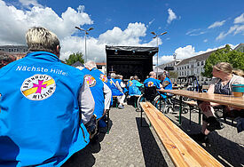 Menschen in blauen Westen mit der Audschrift "Nächste Hilfe Bahnhofsmission" sitzen auf Bänken und blicken zu einer Bühne.