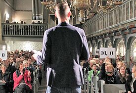 Mann steht vor dem Publikum, man sieht ihn nur von hinten, im Publikum werden Zahlen in die Luft gehalten, alle sitzen in einer Kirche