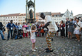 Internationales Jugendnetzwerk-Treffen des Lutherischen Weltbundes: Ein Teilnehmer der Eröffnungsfeier tanzt mit einem kleinen Mädchen auf dem Wittenberger Marktplatz vor dem Luther-Denkmal