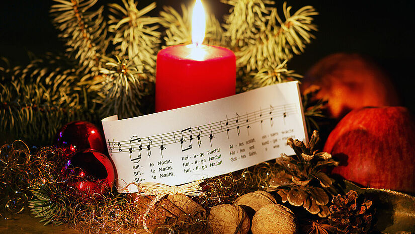 Das Weihnachtslied "Stille Nacht" gehört zu den Klassikern. Einige Chöre werden es in diesem Jahr open air zum Besten geben. Stille Nacht- Ausschnitt auf Papier hübsch dekoriert an weihnachtlicher Deko, Tannenzweige, Nüsse, brennende Kerze, Tannenbaumkugeln