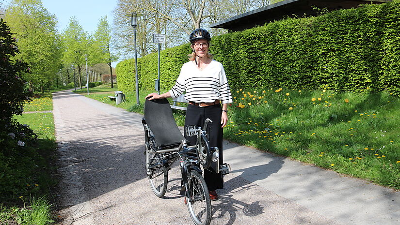 Die Bischöfin trägt einen Fahrradhelm und steht draußen im Grünen neben ihrem Liegerad.