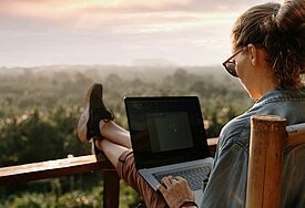 Eine Frau sitzt auf einer Terasse, die Füße lässig auf dem Geländer abgelegt, ein Computer liegt auf ihrem Schoß. Im Hintergrund ist nur Wald, Hügel und Himmel zu sehen.