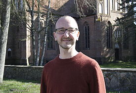 Pastor Felix Degwitz aus der Kirchengemeinde Leussow-Redefin steht in einem roten Pullover vor einer Kirche.