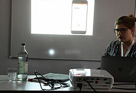 Friederike Nordholt sitzt an einem Tisch, links neben ihr eine per Beamer an die Wand geworfene Folie mit einem Smartphone
