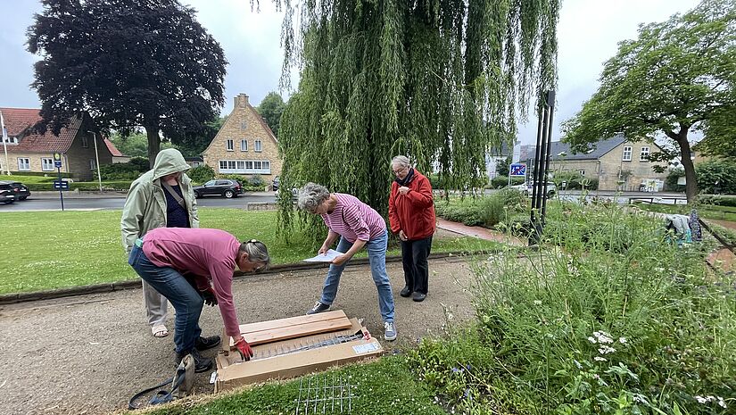 Vier Frauen sind in einer Parkanlage und packen ein längliches Paket mit einer Gartenbank aus. 